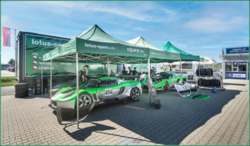 Vozy KT Motorsport pod stany lotus-sport.com v padoku, mechanici u zeleného vozu 494.