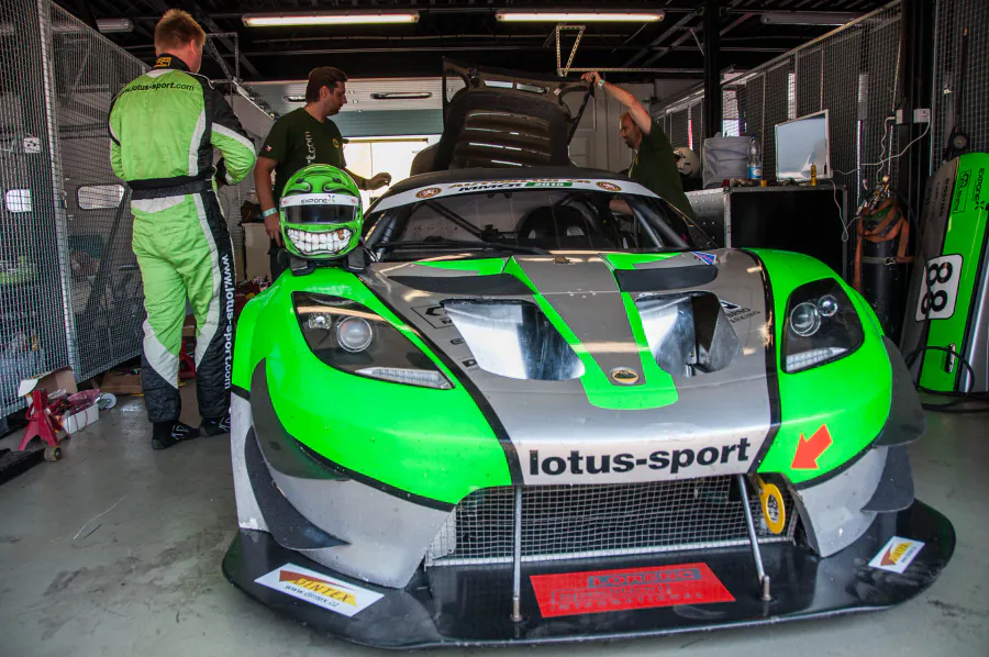Zelený závodní Lotus Exige otevřený v boxu, dva mechanici u přední části vozu.
