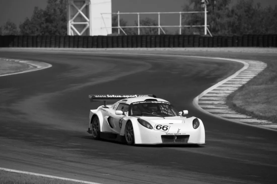 Bílý závodní Lotus Exige se startovním číslem 66 a nápisem lotus-sport.com projíždí zatáčkou okruhu, černobílá archivní fotografie.